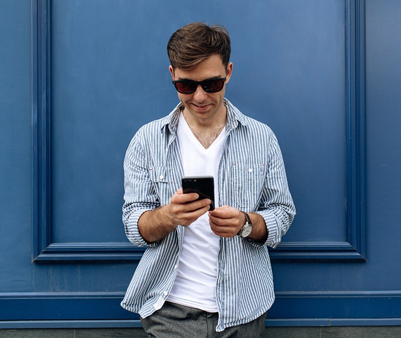 A young man in sunglasses checks his smartphone, researching how to delete his Facebook account.