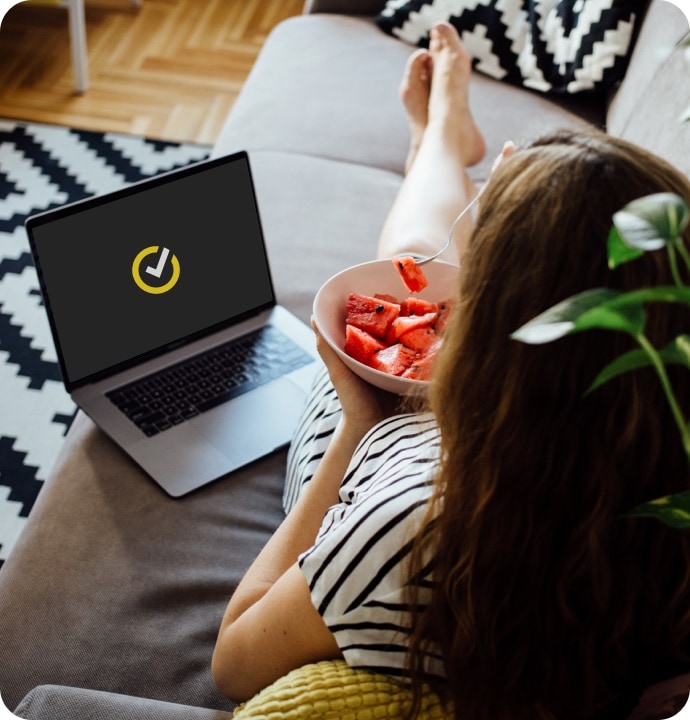 Woman lying on a couch eating a bowl of fruit with a laptop beside her displaying Norton logo